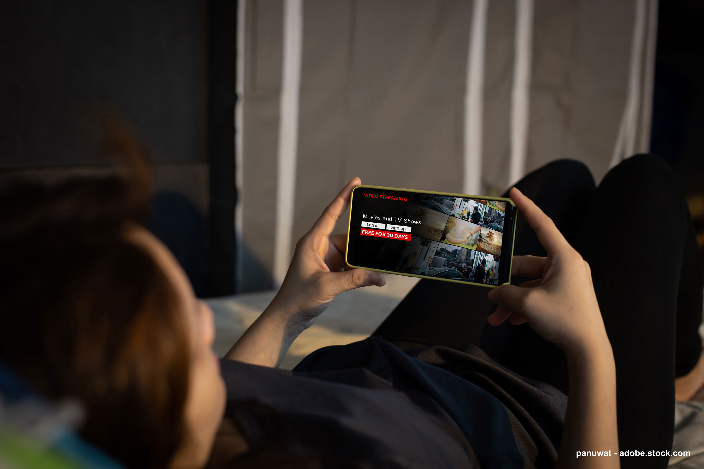 Un jeune est allongé sur son lit et regarde, sur son téléphone, du contenu audiovisuel soumis à un abonnement