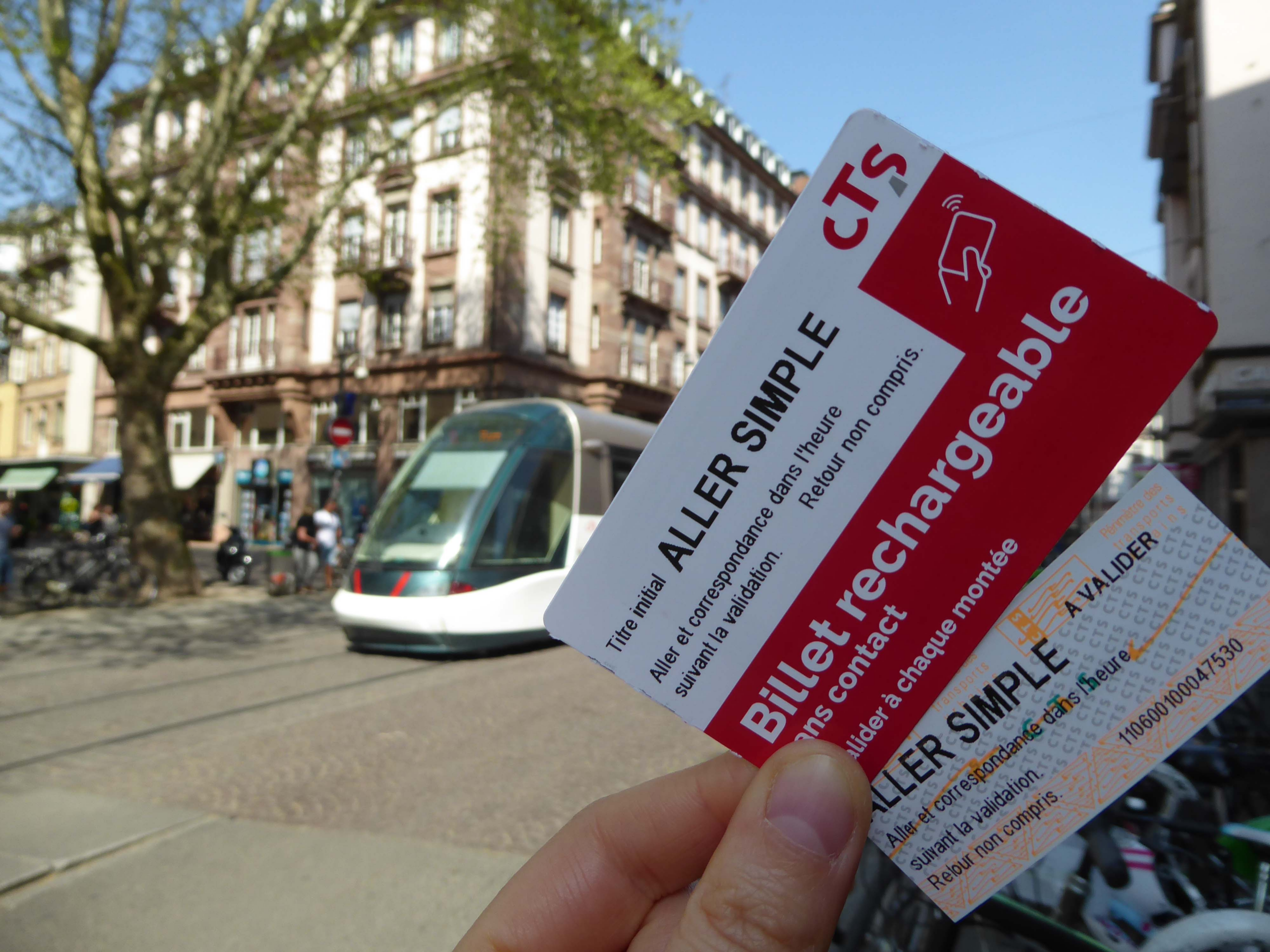 Eine Hand hält zwei Fahrkarten des Straßburger Nachverkehrsunternehmens CTS, im Hintergrund fährt eine Tram.