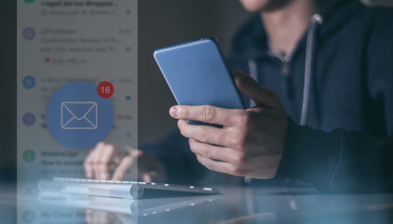 Mann hält ein Smartphone, tippt auf einer Tastatur und liest E-Mails, im Vordergrund ein E-Mail-Icon