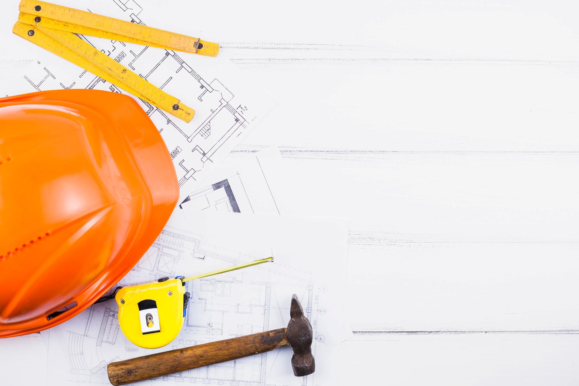 Auf Plänen mit Grundrissen liegen ein Zollstock, ein orangener Baustellenhelm, ein Bandmeter und ein Hammer.