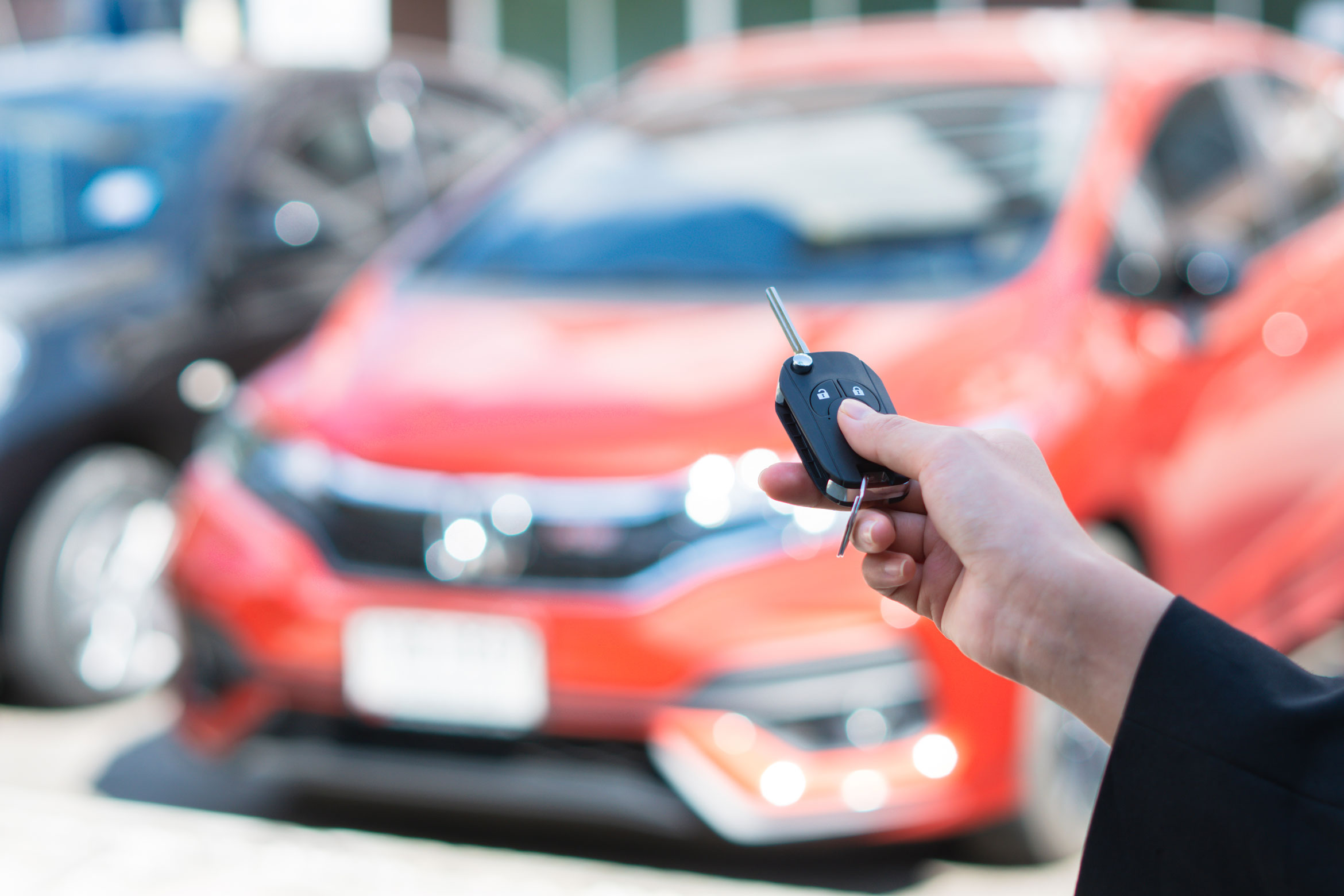 Une main tenant une clé de voiture à l'avant-plan, et une voiture rouge à l'arrière-plan