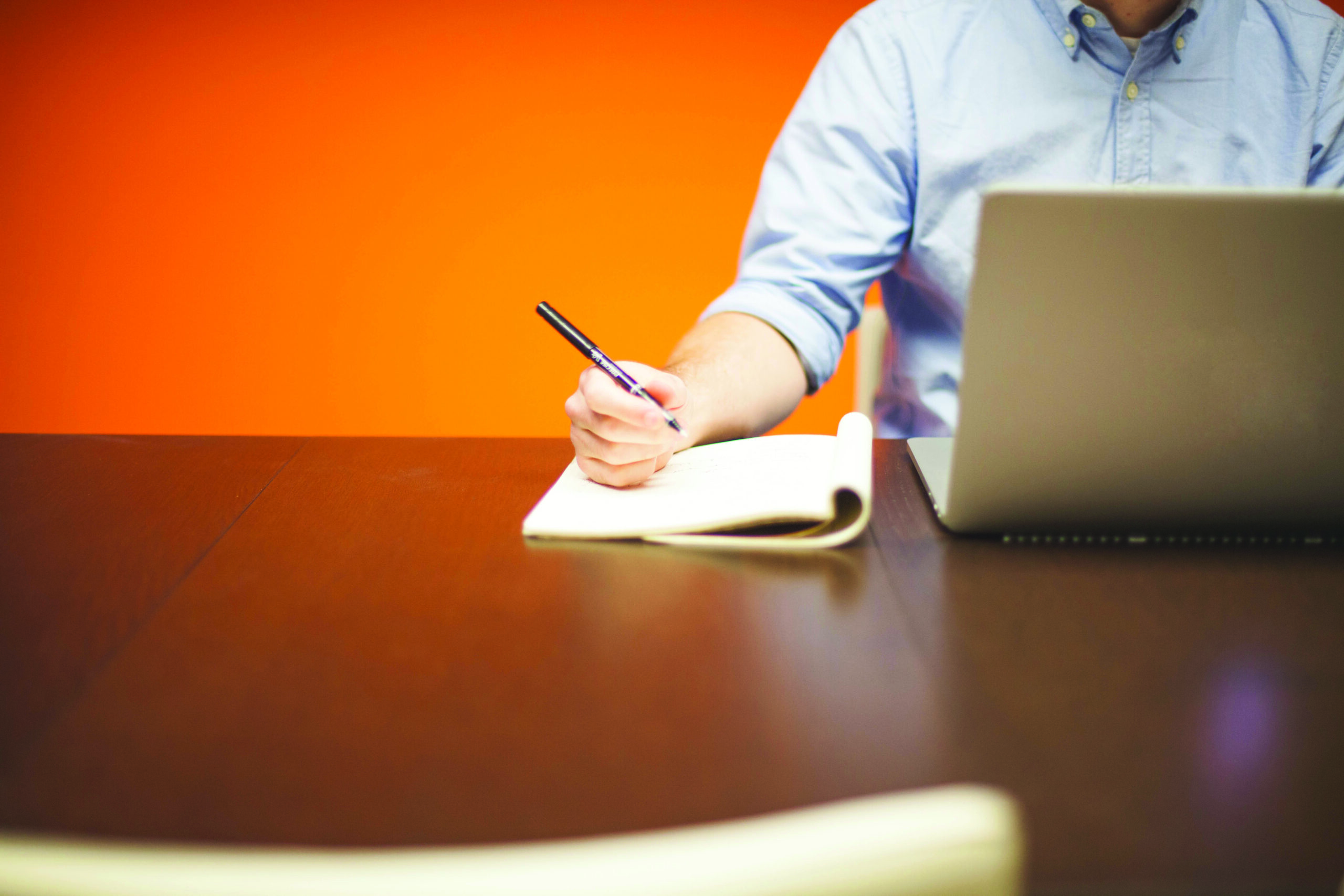 Un homme assis devant un ordinateur portable et un cahier, le stylo à la main.