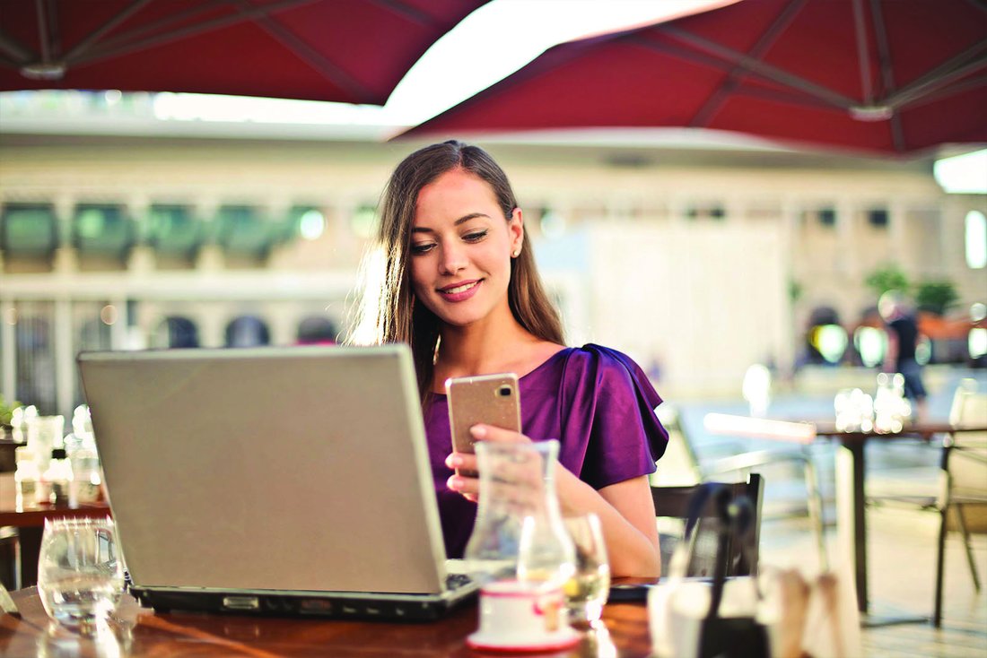 Une femme assise en terrasse consulte son téléphone ainsi que l'ordinateur portable posé devant elle sur la table.