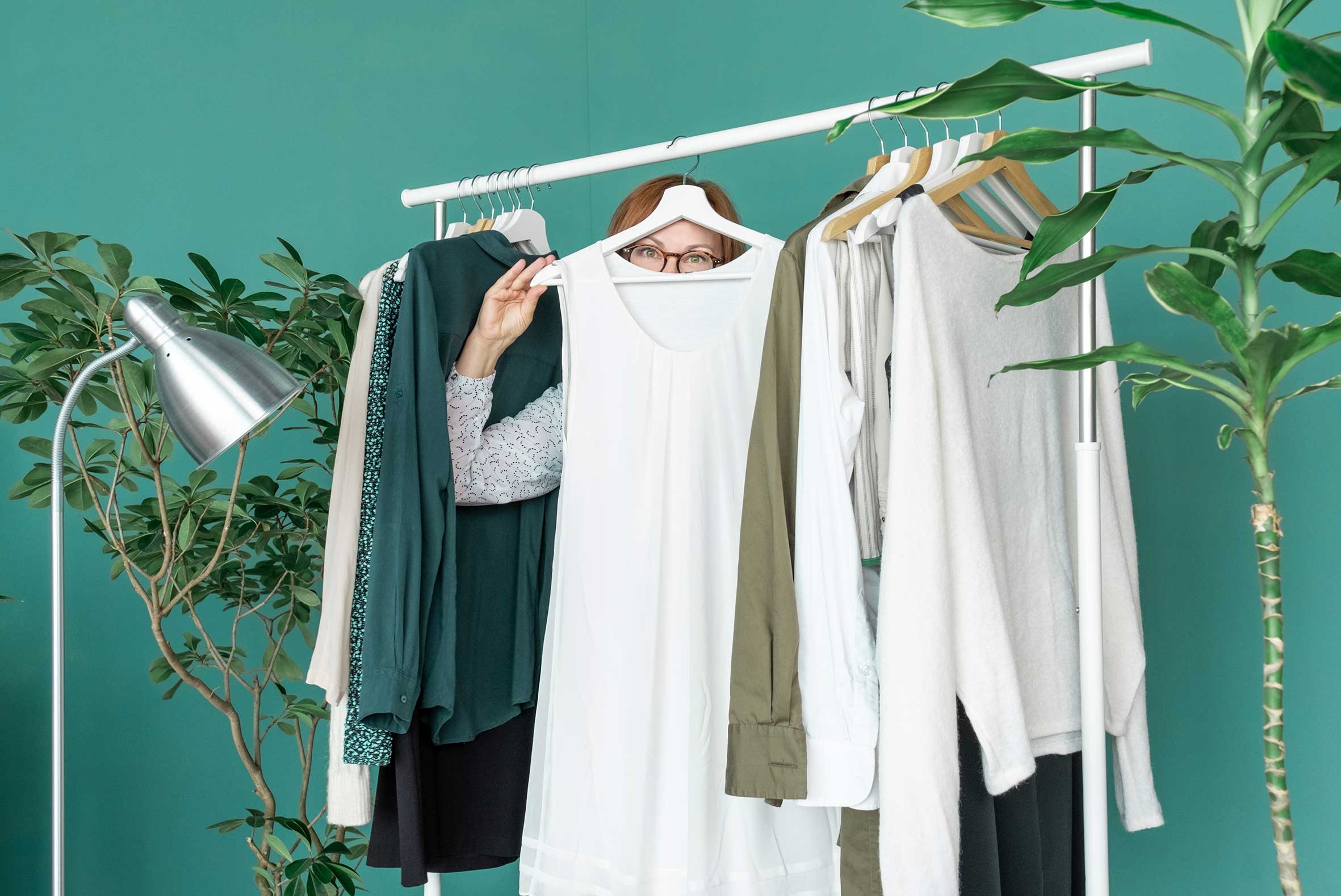 Eine Frau versteckt sich hinter einem weißen Kleid, das an einer Kleiderstange hängt. Daran hängen noch weitere Kleidungsstücke und Pflanzen stehen daneben.