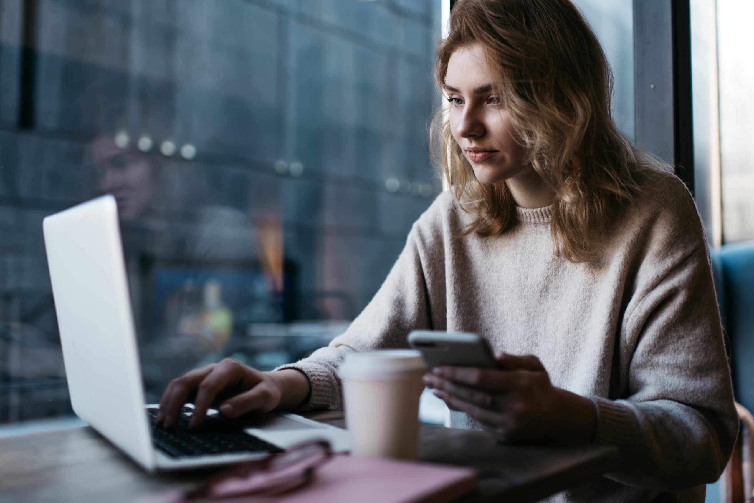 Eine Frau sitzt in einem Café und arbeitet an einem Laptop und hält ihr Smartphone in der Hand.