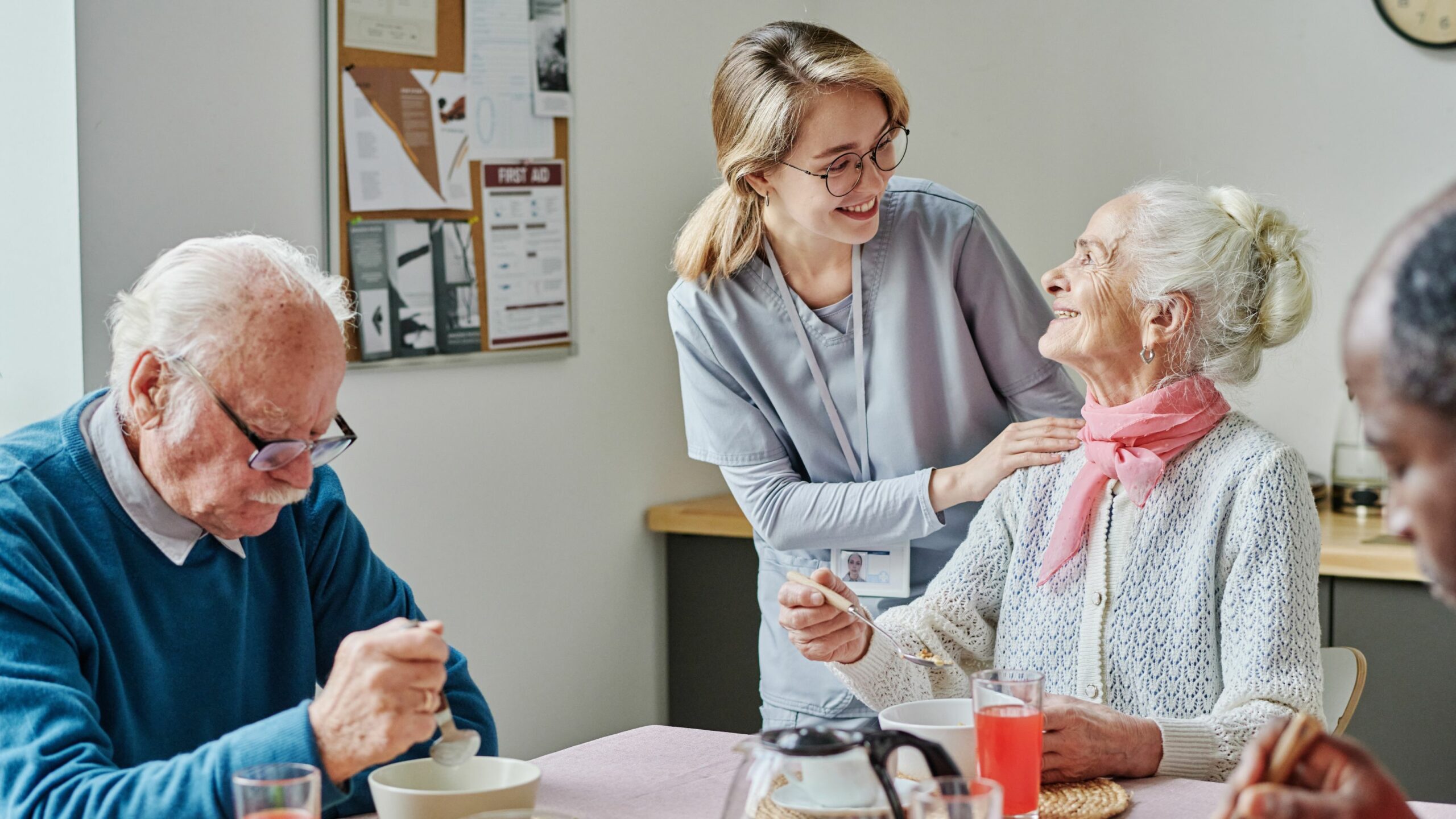 Ein Senior und eine Seniorin sitzen am Frühstückstisch und sprechen mit der Pflegerin.